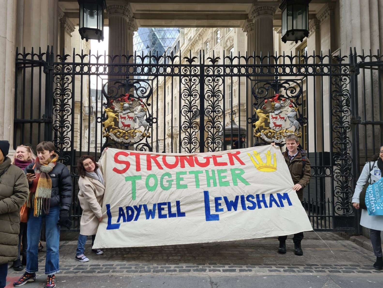 Photo of Protect Prendergast protesters who are outside closed front gate to the Leathersellers Hall in London holding banner which says “Stronger Together Ladywell Lewisham”